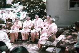 Multiple Swamis seated with food on little tables in the garden, devotees in background